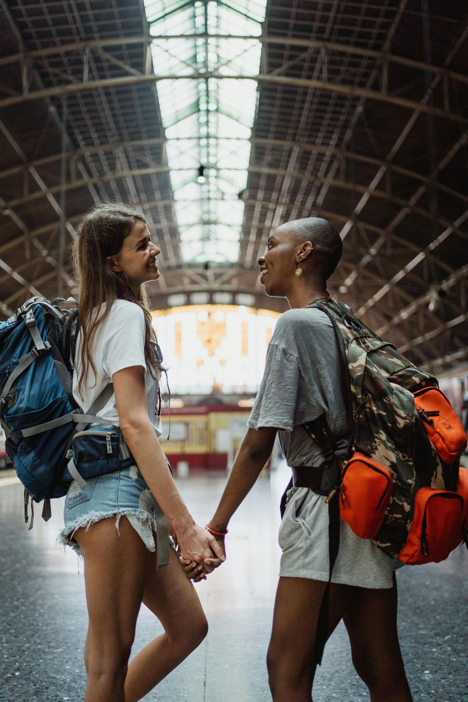 Two women holding hands in a train station, smiling at each other. They wear backpacks and casual clothes.