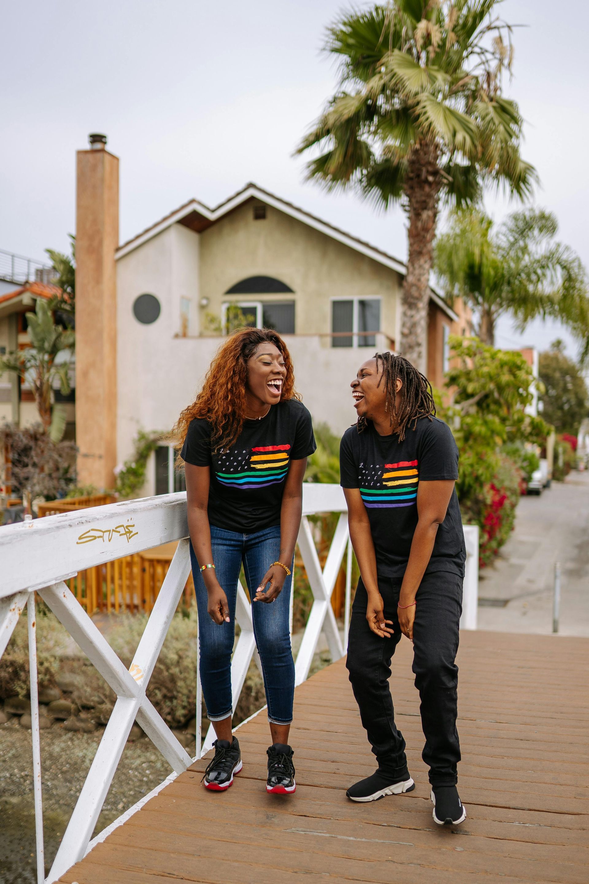Two laughing Black women on a bridge wearing matching rainbow shirts, palm trees and houses in the background.
