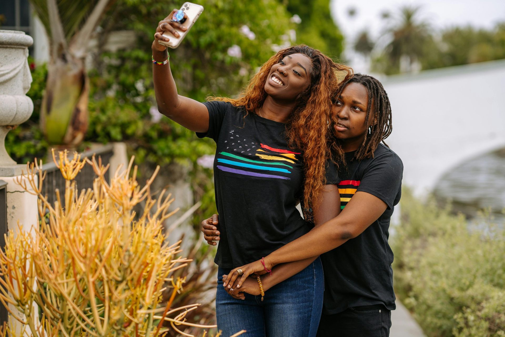Two people taking a selfie. One person holds the phone, smiling. The other hugs them, with a neutral expression. Both wear matching pride shirts.
