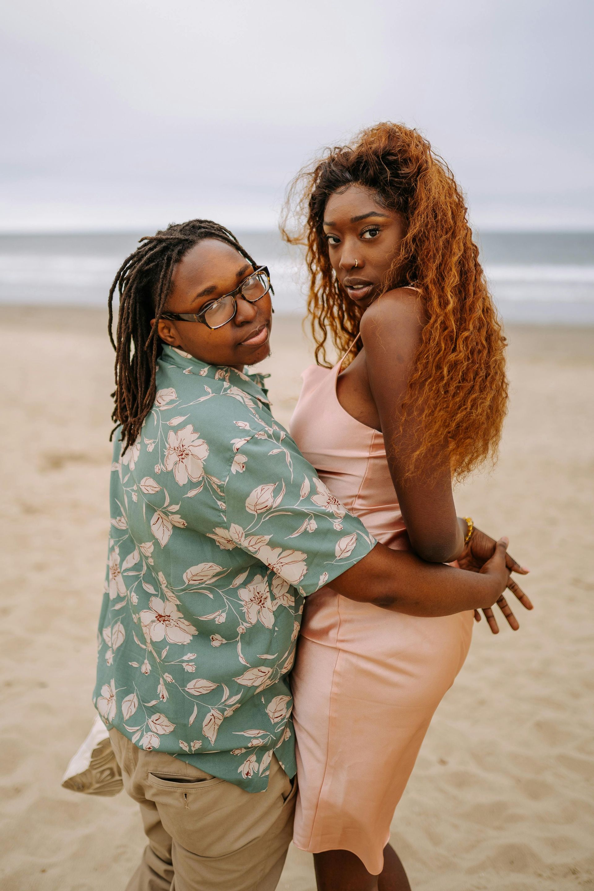 Two people embracing on a beach; one in a floral shirt, the other in a pink dress, looking at the camera.