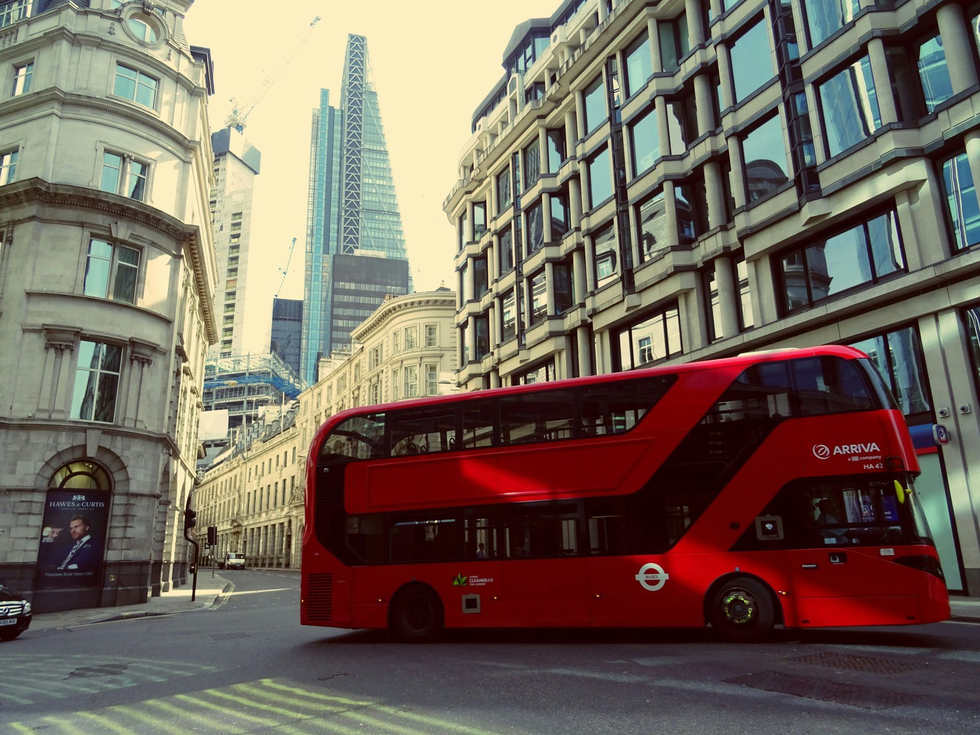 A red double-decker bus drives through a narrow London street surrounded by tall stone buildings and a modern skyscraper.