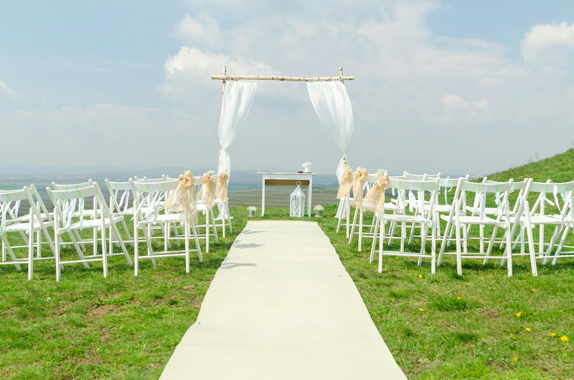 Wedding ceremony setup on a grassy hilltop with white chairs, aisle runner, and draped arch.