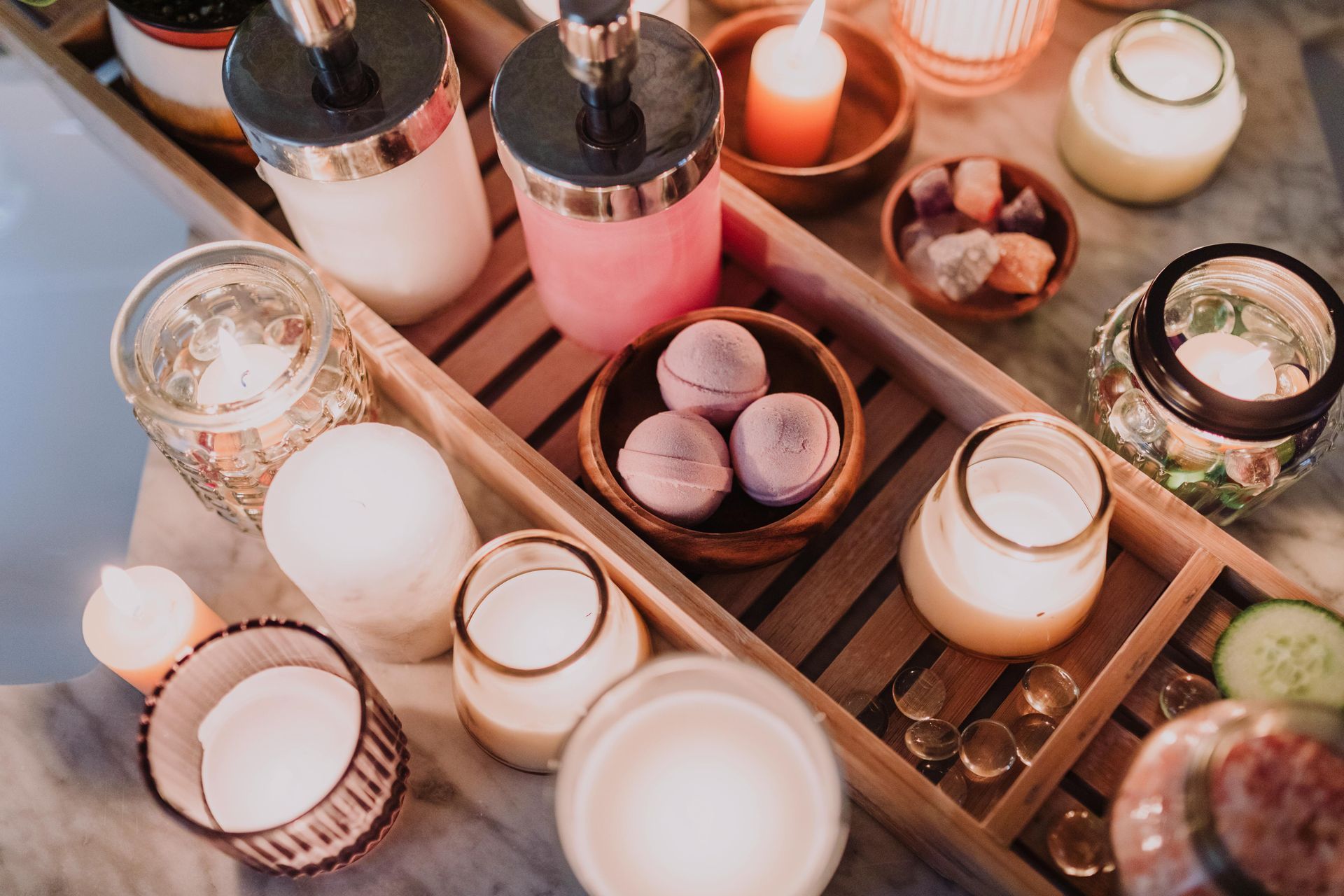 A wooden bathtub tray holds soap dispensers, candles, bath bombs, and small bowls filled with colorful crystals.