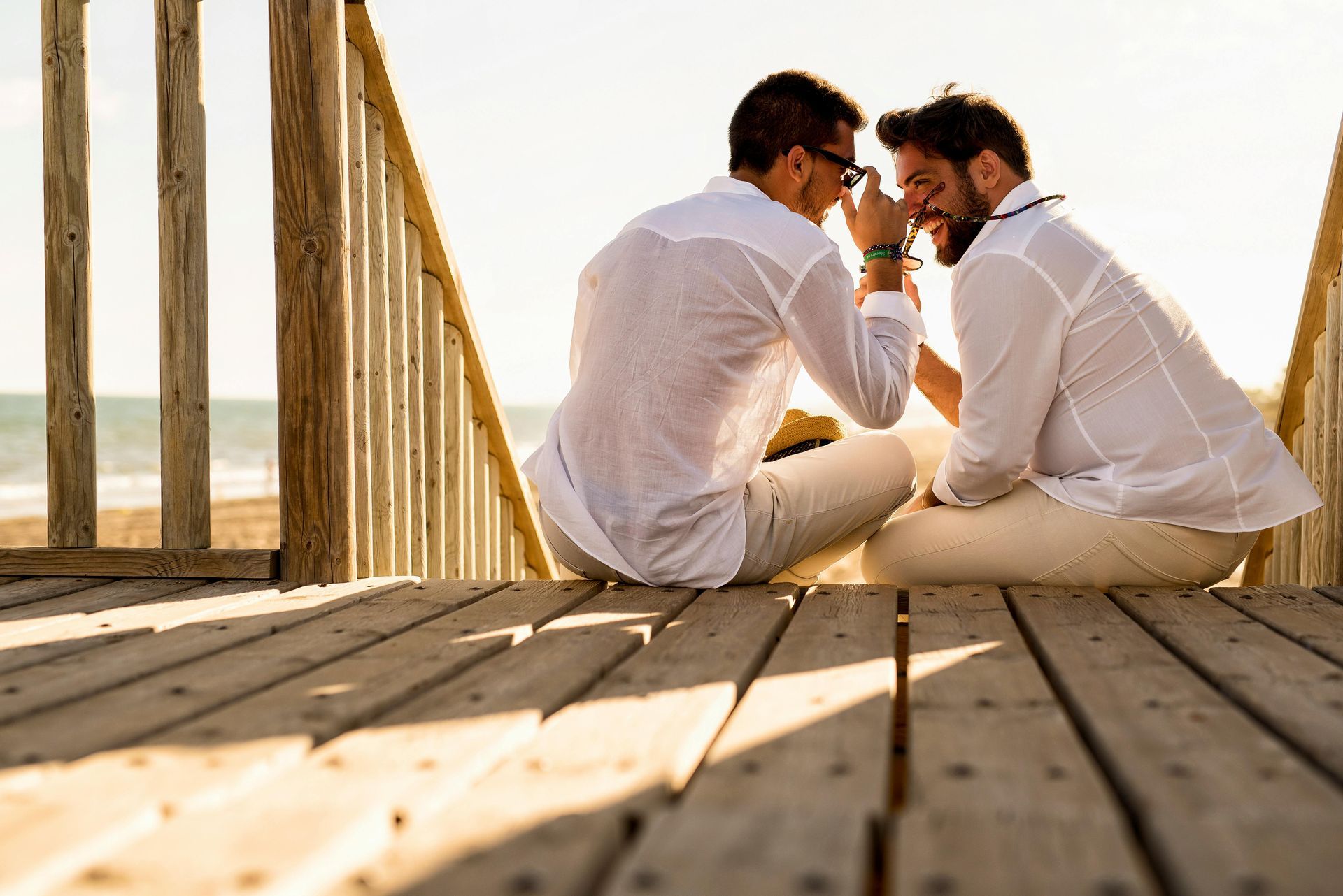 Two men in white shirts and pants sit close together on a wooden walkway at the beach.