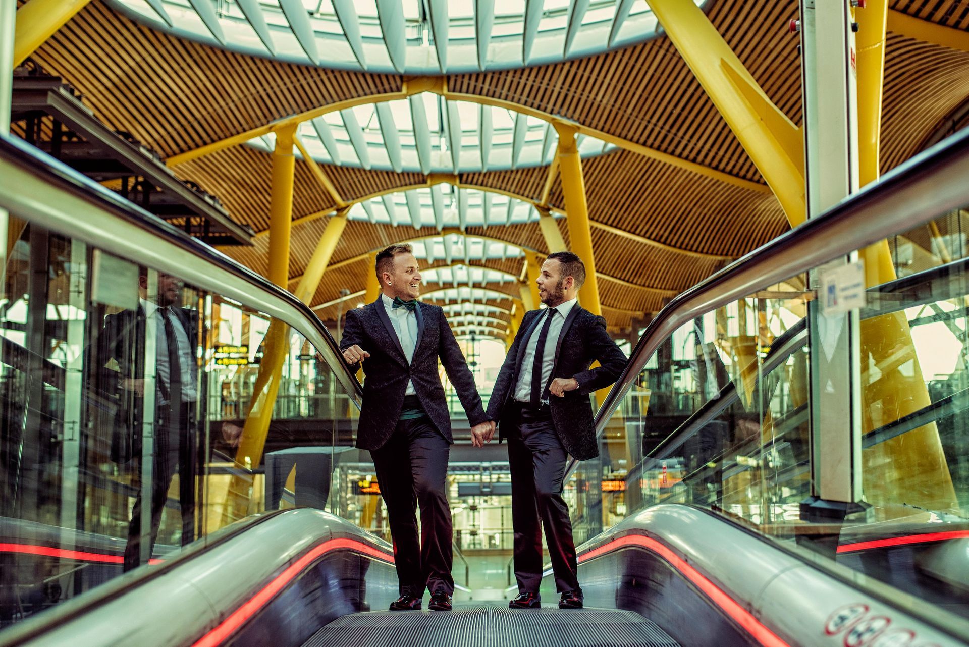 Two men in suits holding hands, on escalator in modern building with high ceiling.