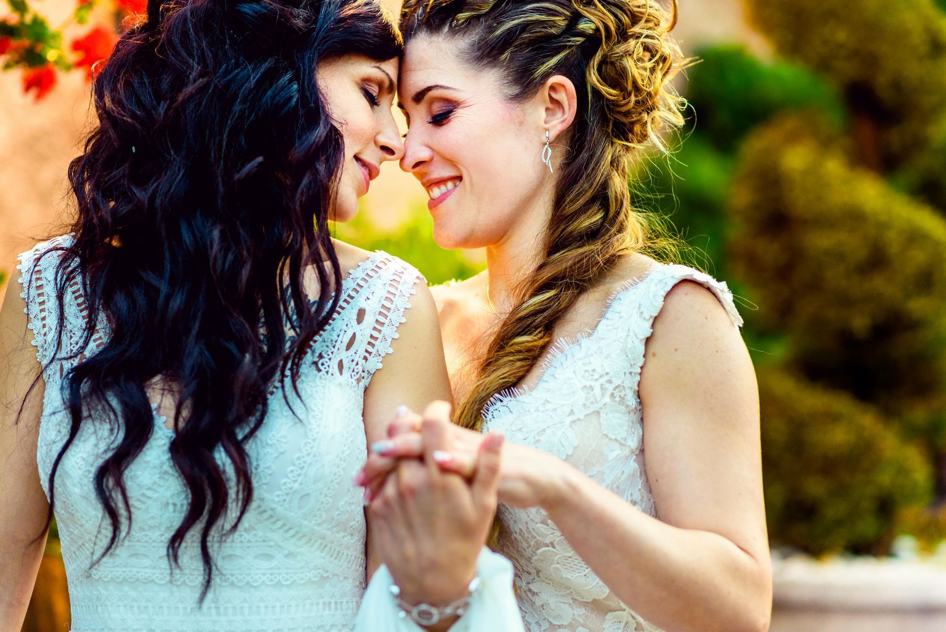 Two people in white wedding dresses hold hands and touch foreheads, smiling softly in an outdoor garden setting.