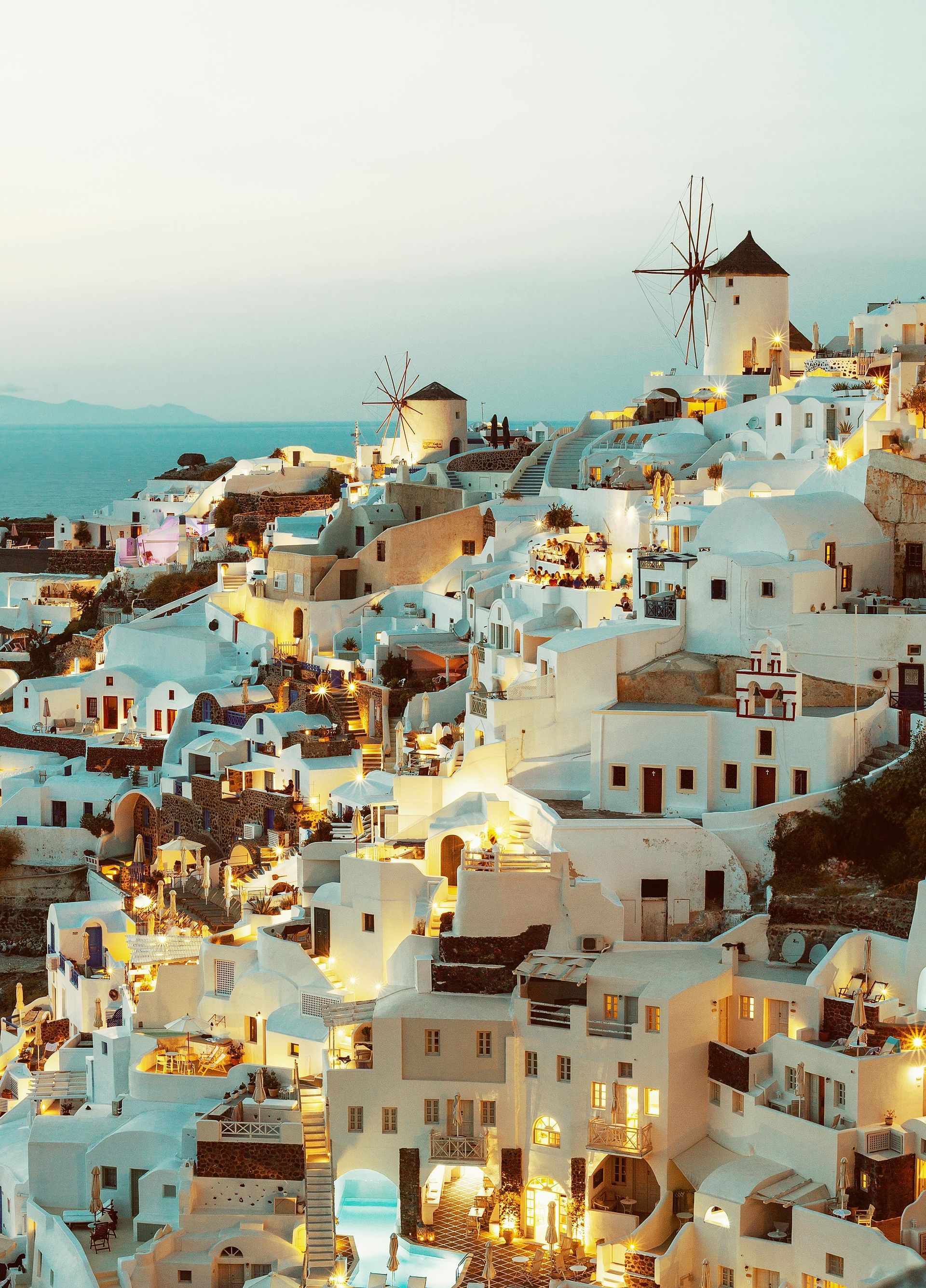 A high-angle view of Oia, Santorini, featuring iconic white buildings and windmills glowing under a soft twilight sky.
