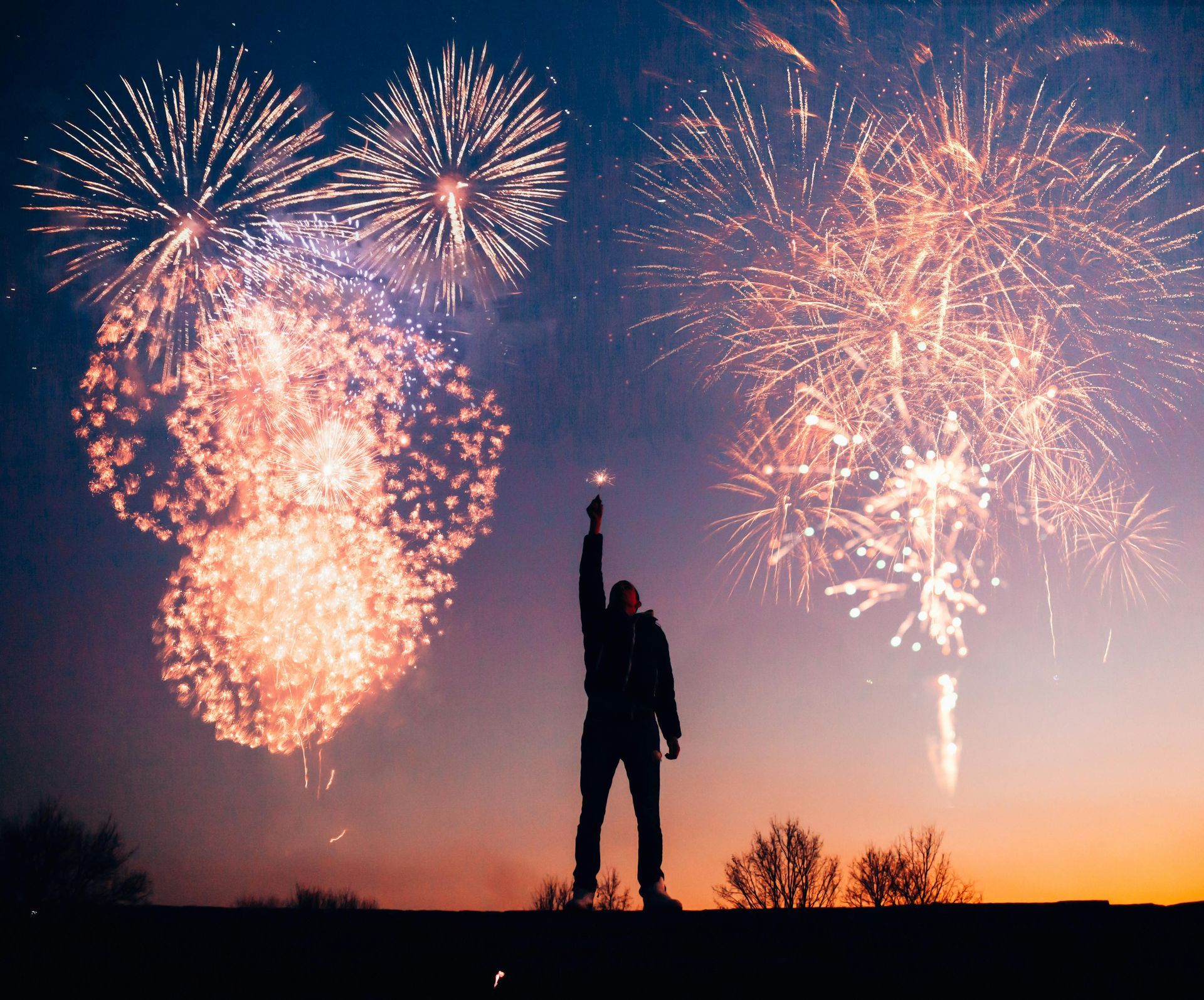 Silhouette of person with raised arm holding sparkler, watching fireworks against sunset sky.