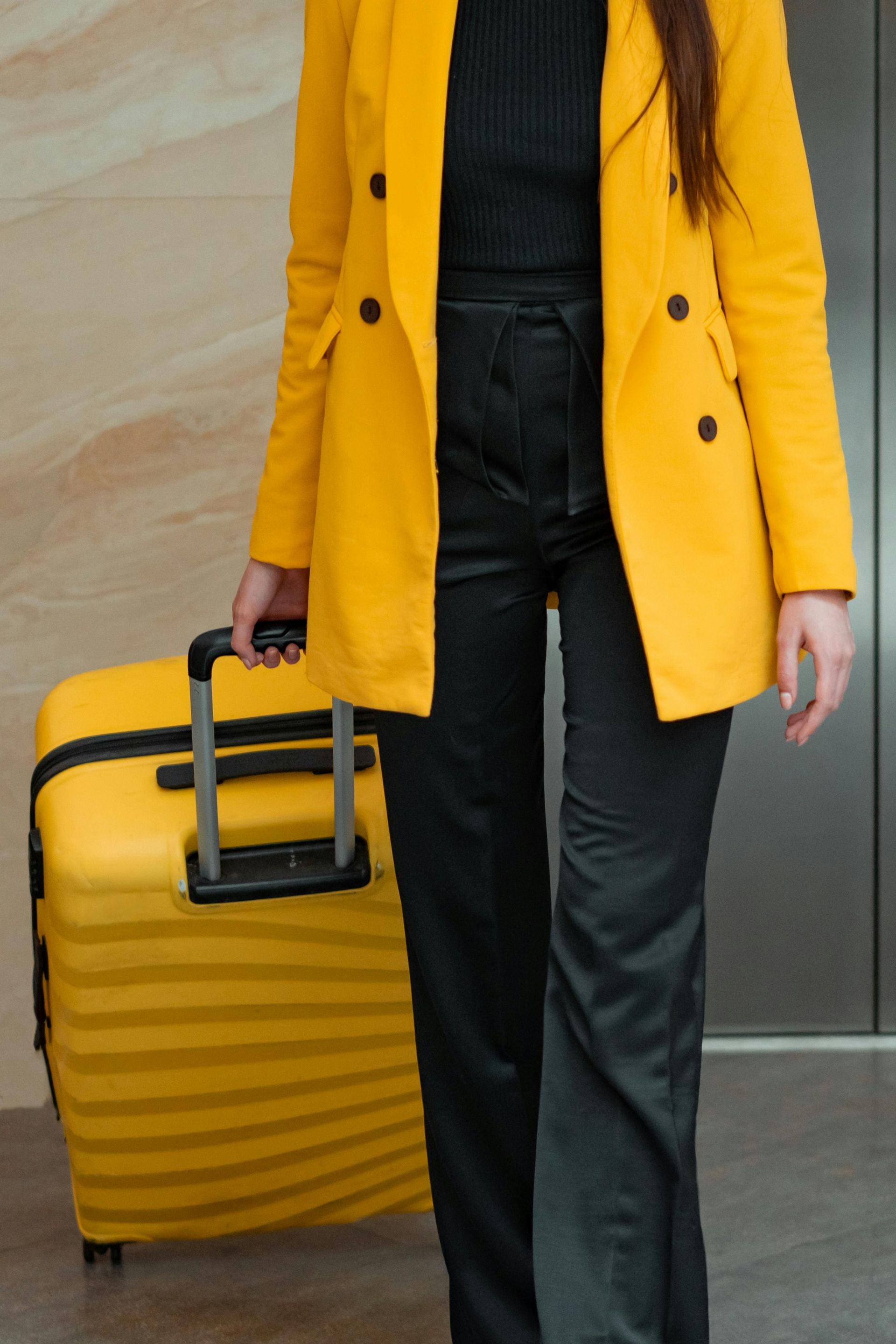 A person in a yellow blazer and black trousers walks while pulling a matching yellow rolling suitcase.
