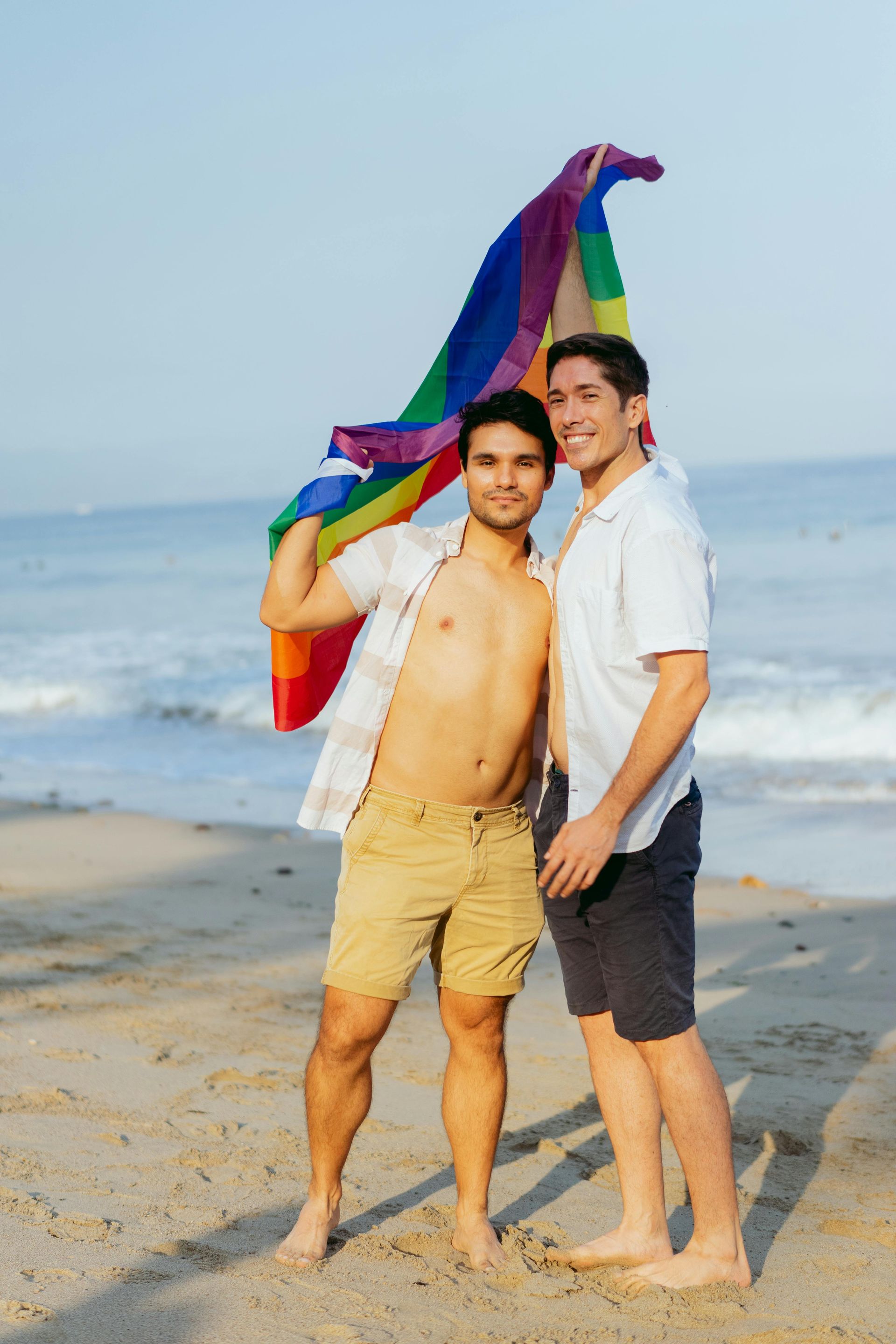 Two people on a beach hold a rainbow flag. One shirtless, smiling. Sunny day, ocean in the background. Phuket