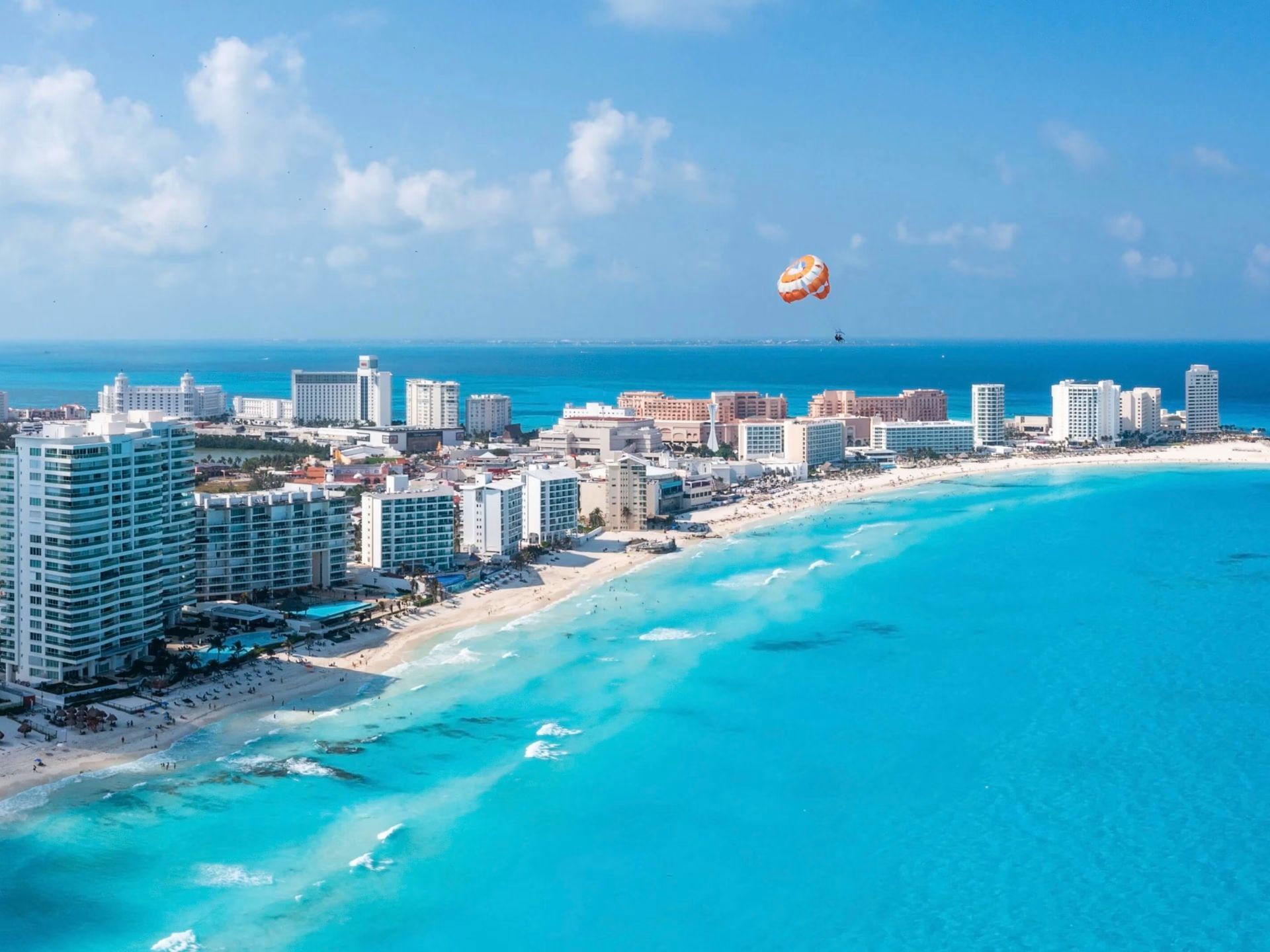 Coastal cityscape with turquoise water and white sand beach, parasailer overhead, sunny day. Cancun