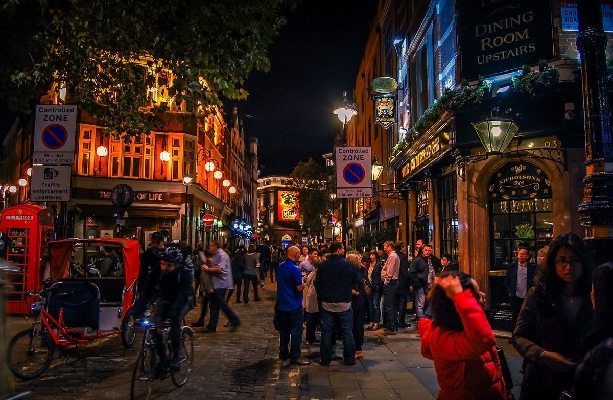 Nighttime street scene with illuminated buildings, crowds, cyclists, and a red phone booth.