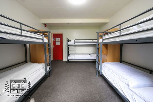 Dorm room with four bunk beds, white bedding, carpeted floor, and a red door at the back.