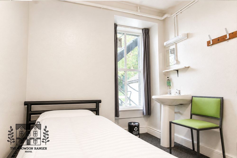 Small bedroom with white walls, twin bed, window, shelf, and chair by a corner sink.
