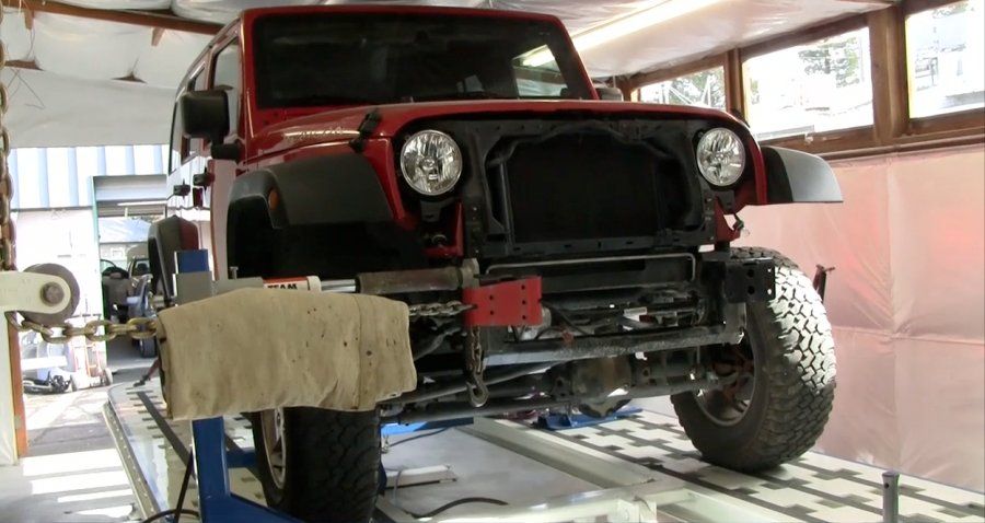 Red Jeep Wrangler on a lift in a garage, front grill removed, wheel off.