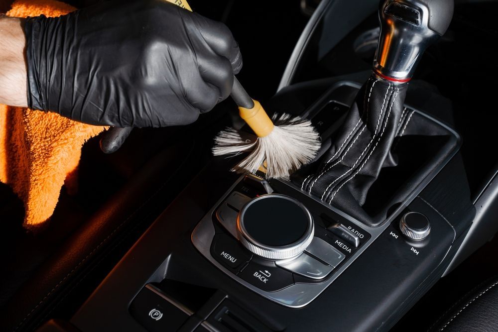 Person in black gloves cleaning car console with a brush. Orange cloth visible.