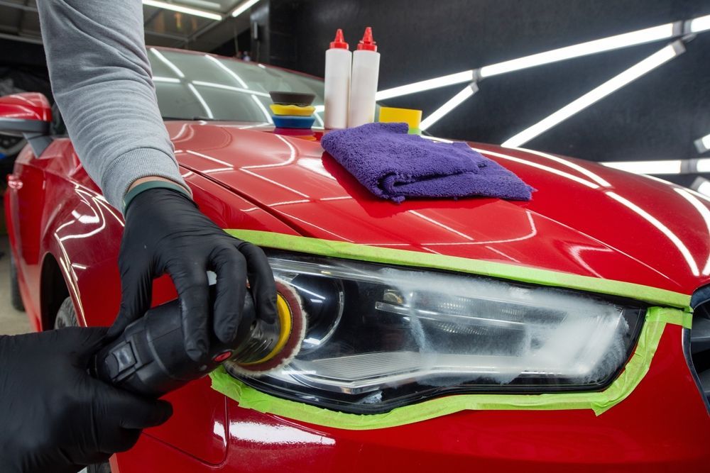 Person polishing car headlight with a power tool, red car, black gloves, green tape.