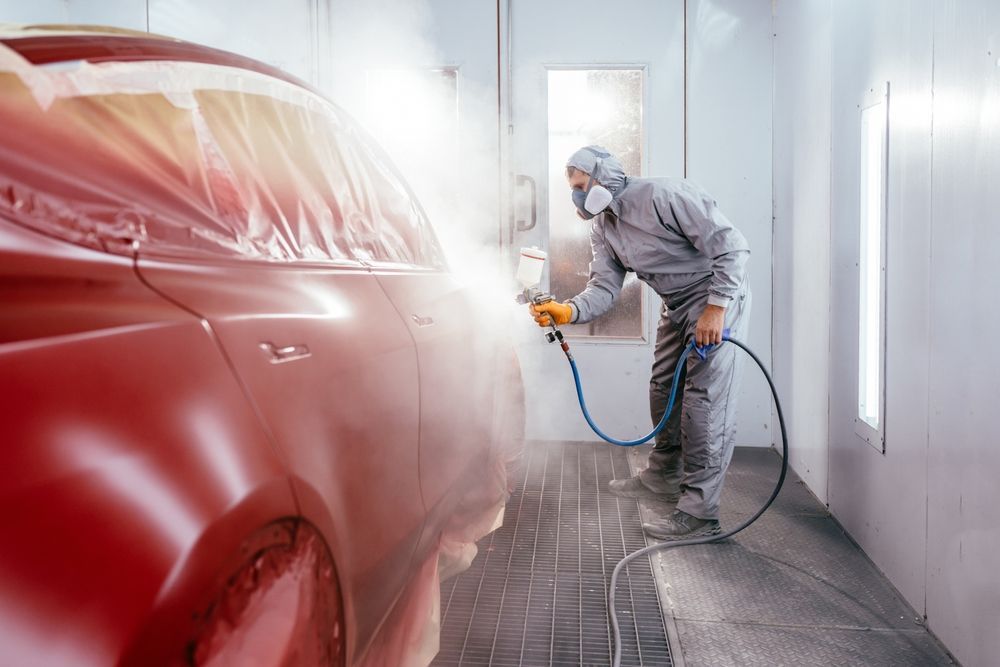 Person in protective suit spraying red paint onto a car in a paint booth.