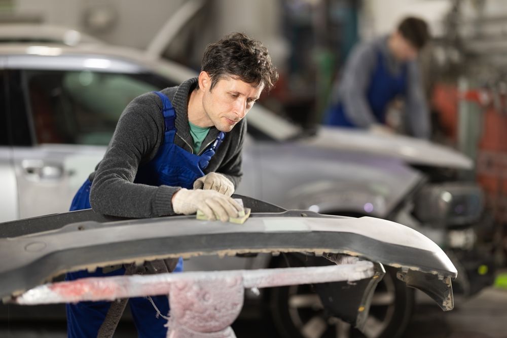 Mechanic sanding a car bumper in a garage, another person in the background.