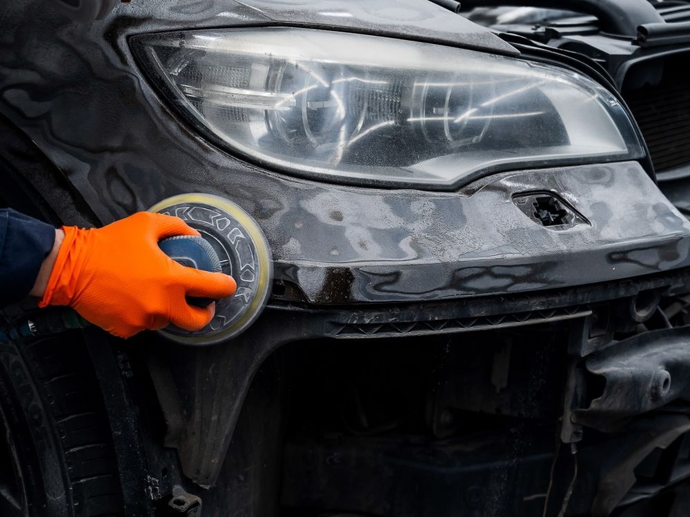 A person in orange gloves uses a sander on a car's black bumper near the headlight.