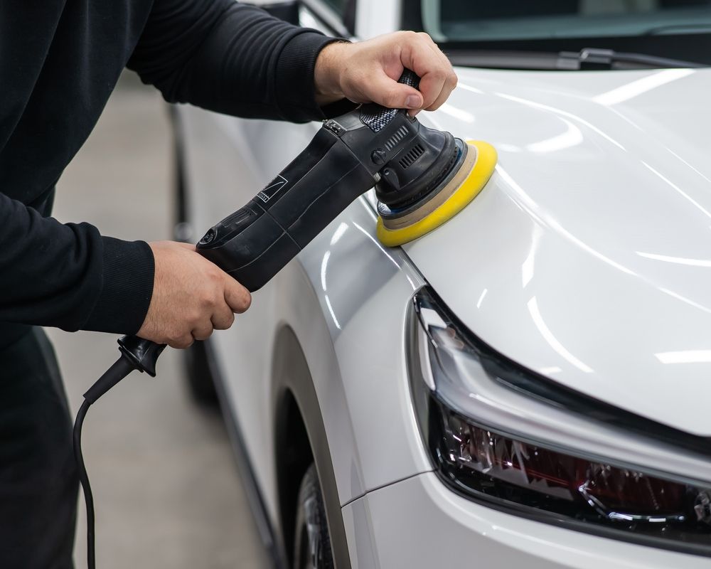 Person polishing a white car hood with an electric buffer in a well-lit shop.