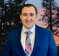Man in blue suit and tie, smiling at the camera; forest background.