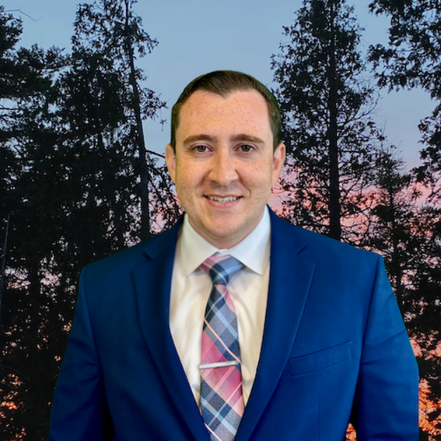 Man in blue suit and patterned tie smiling, with a forest background.
