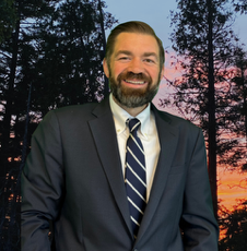 Man in a suit smiling, with a forest background.