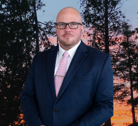 Bald man in navy suit, pink tie, and glasses, set against a sunset with trees.