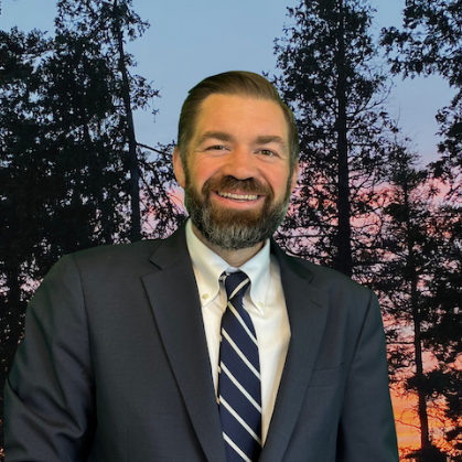 Man in suit, smiling, against a forest backdrop at sunset.