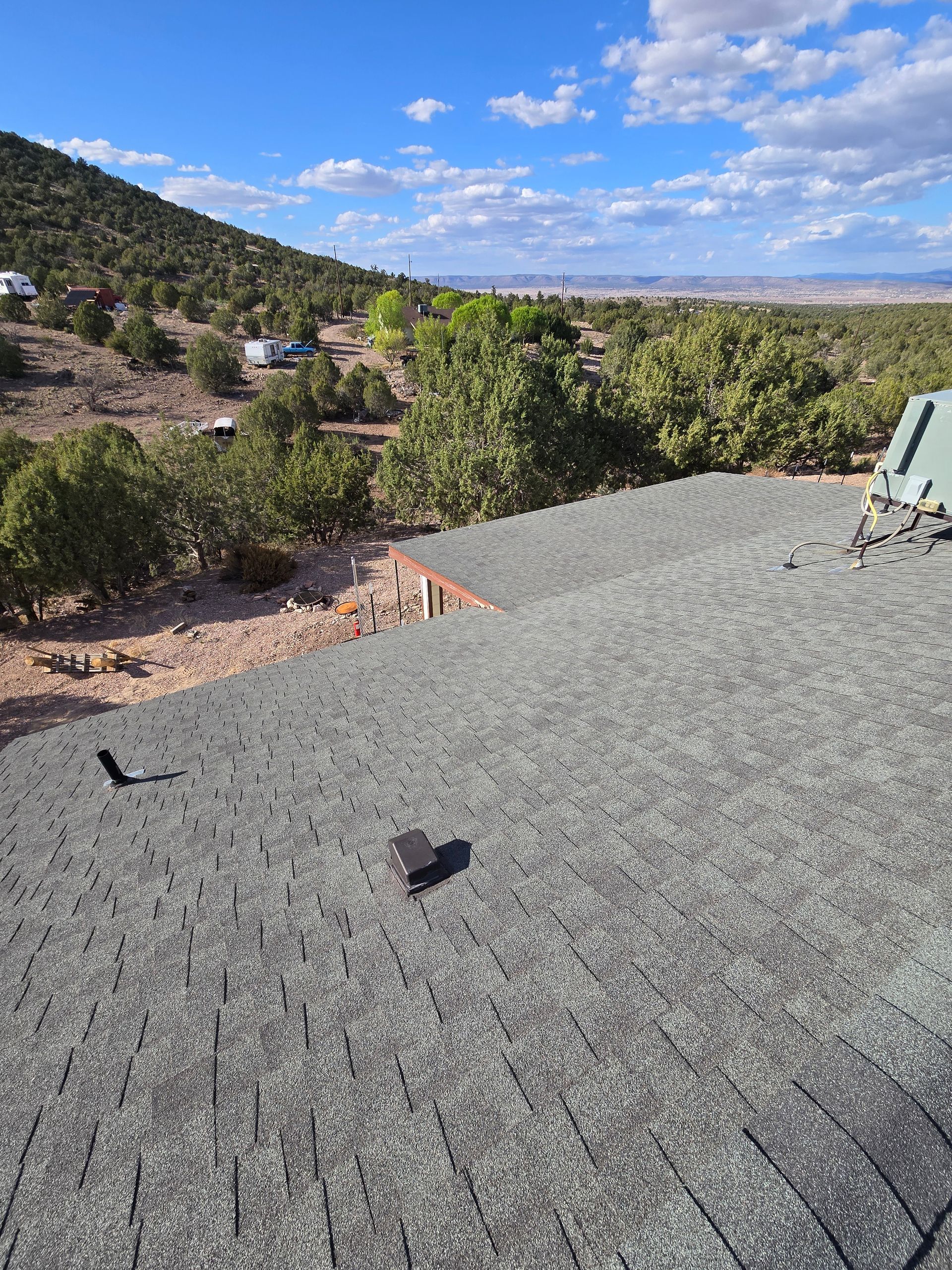 View from a rooftop: grey asphalt shingles, trees, and a mountain range under a blue sky with clouds.