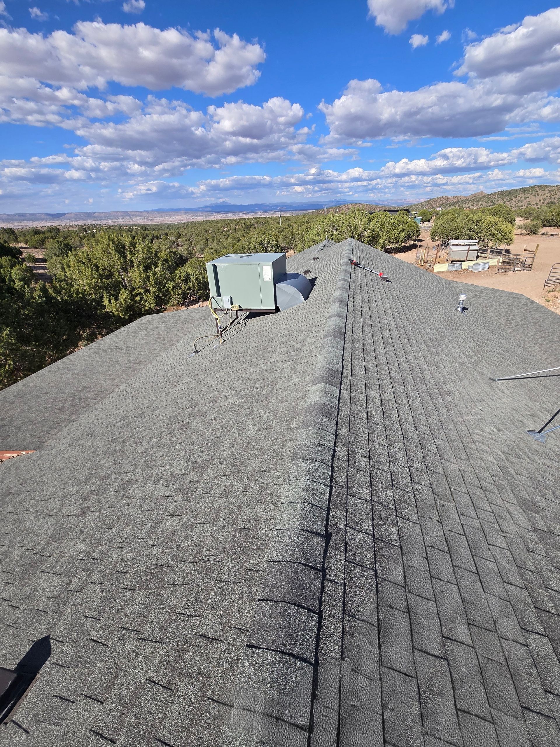 Roof with gray asphalt shingles, blue sky with clouds in the background. An HVAC unit sits on the roof.