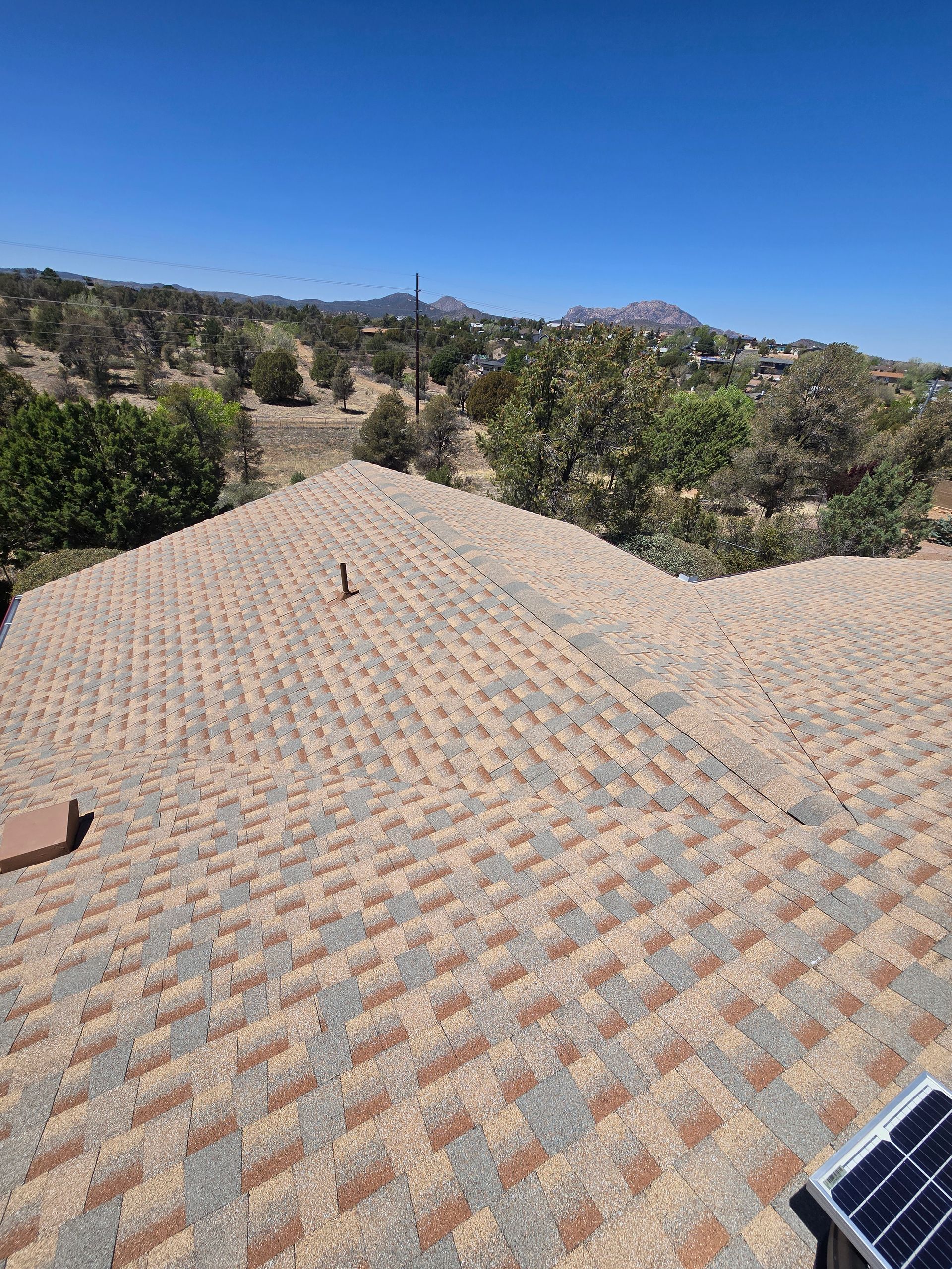 View of a shingled roof with a clear blue sky and a landscape of trees and hills in the background.