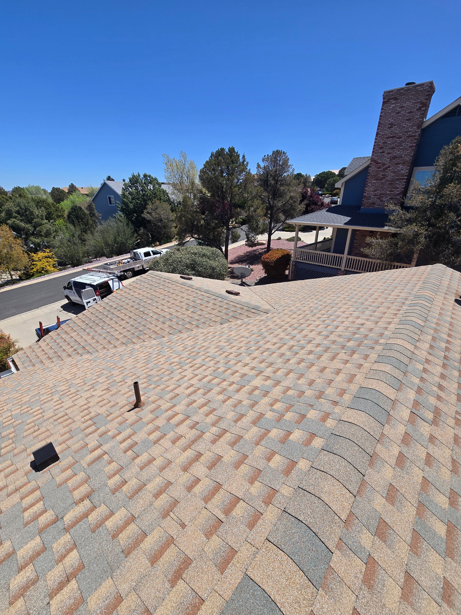 View of a shingled roof with a chimney, houses, and trees under a clear blue sky.
