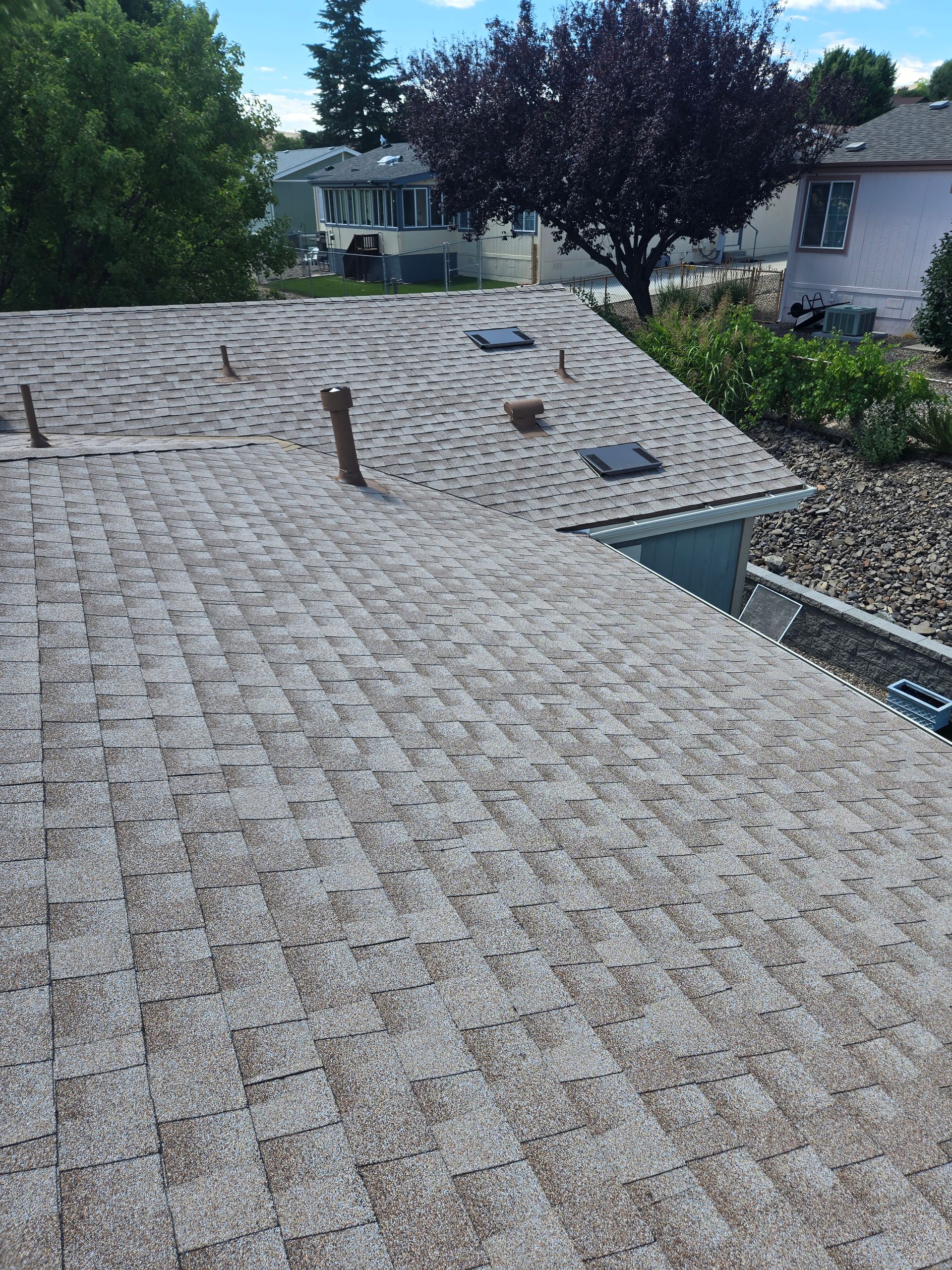 Gray asphalt shingle roof on a house, with several vents and a tree in the background under a blue sky.