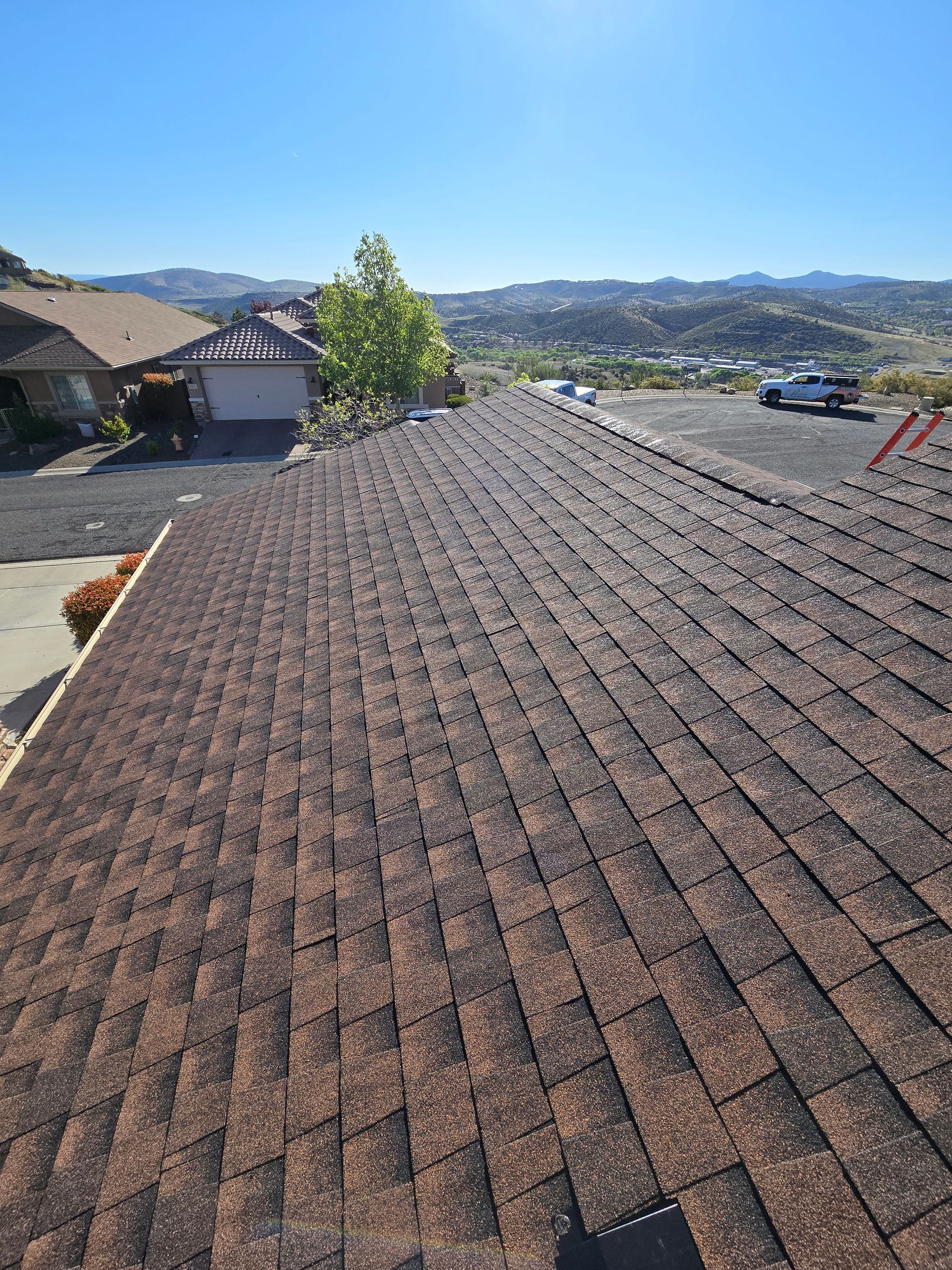 View from a shingled roof overlooking a sunny suburban neighborhood and distant hills