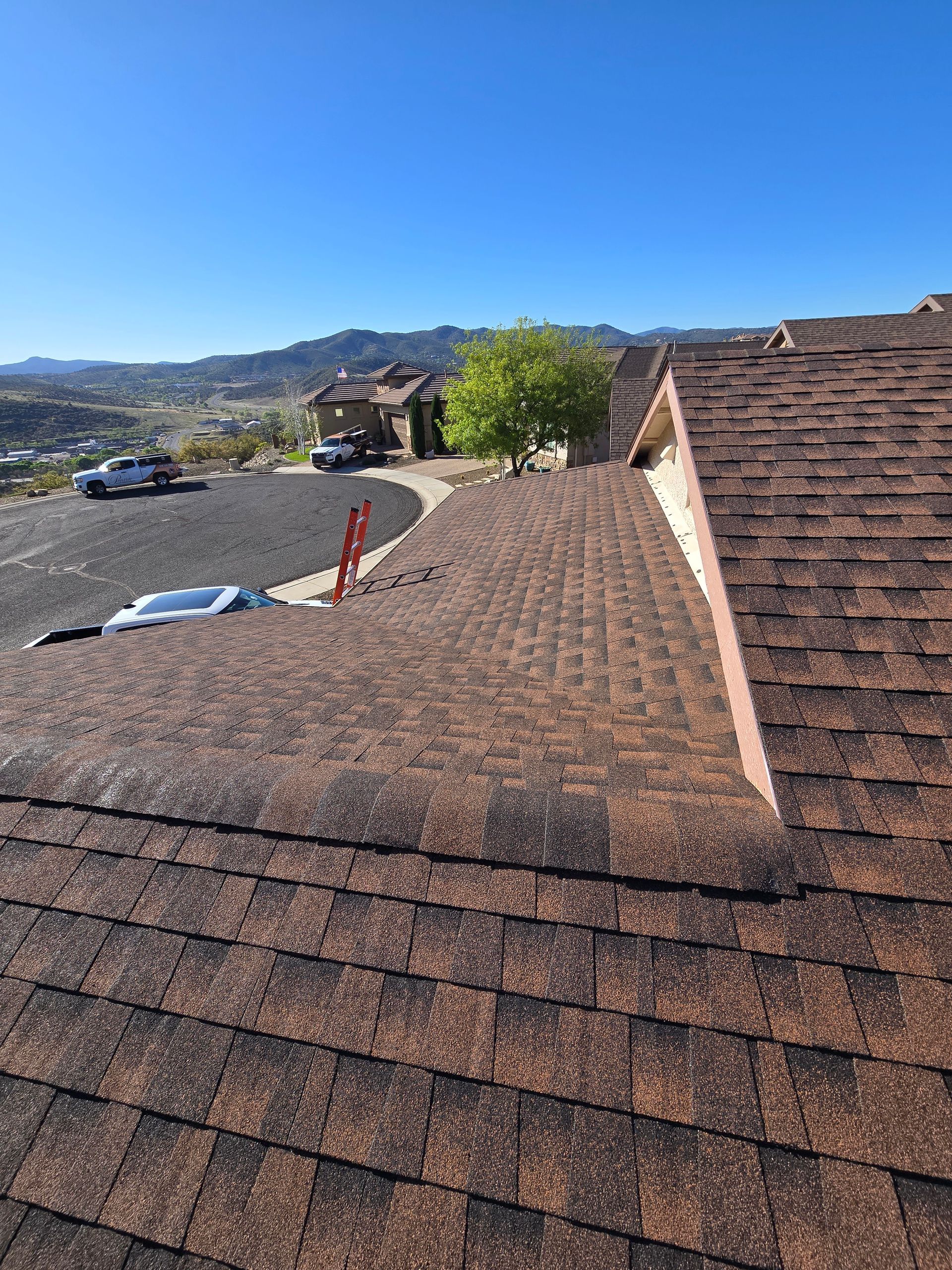 Roof shingles on a house overlooking a sunny hillside parking lot and mountains under a clear blue sky