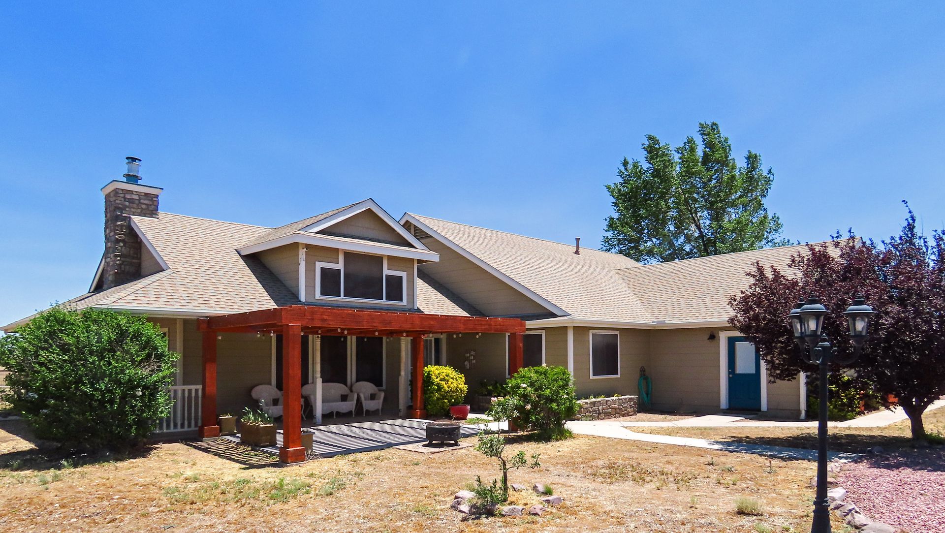 Single-story suburban house with tan roof, red porch posts, and dry front yard under a blue sky