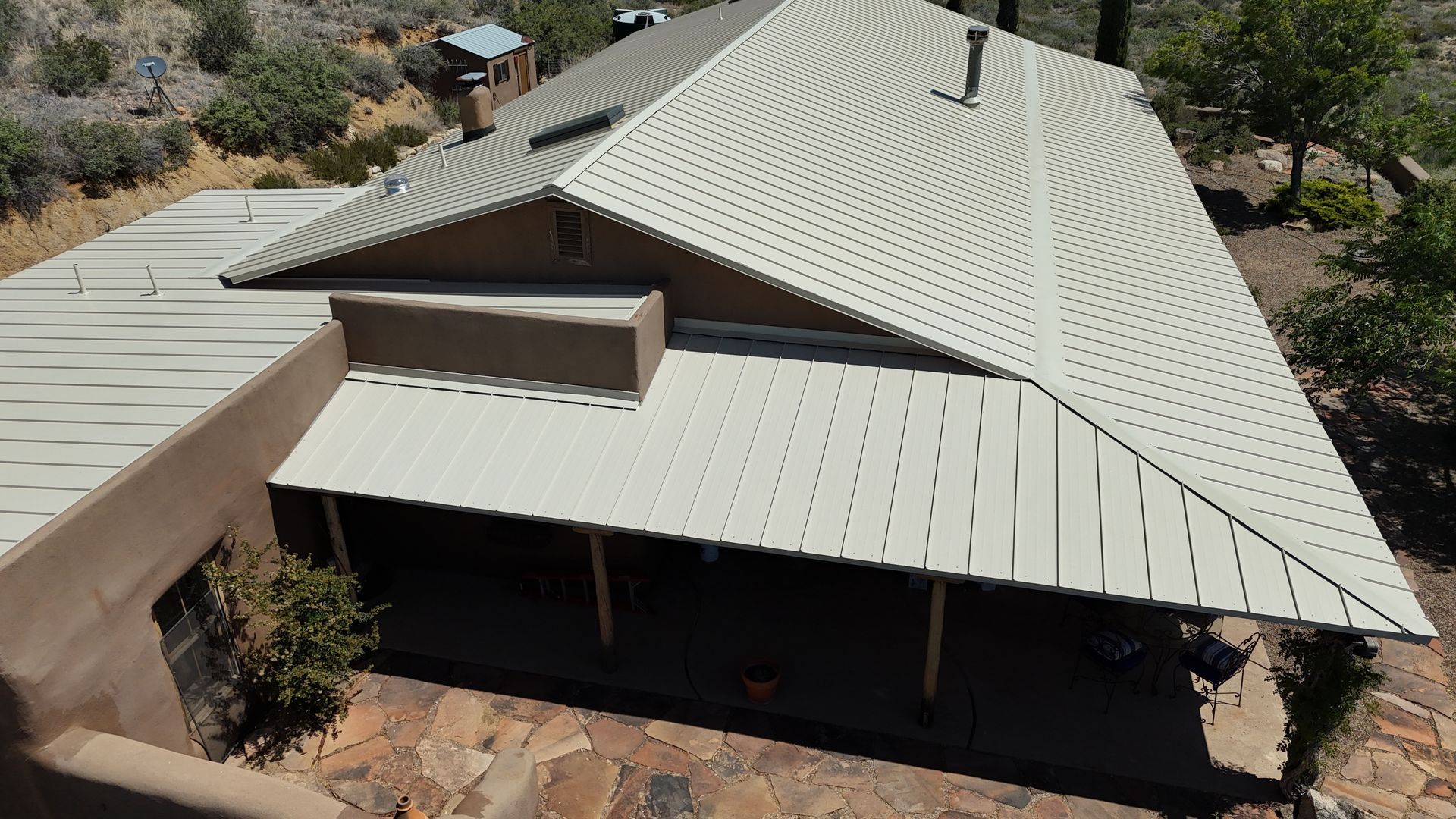 Aerial view of a beige metal-roof house surrounded by trees and rocky ground.