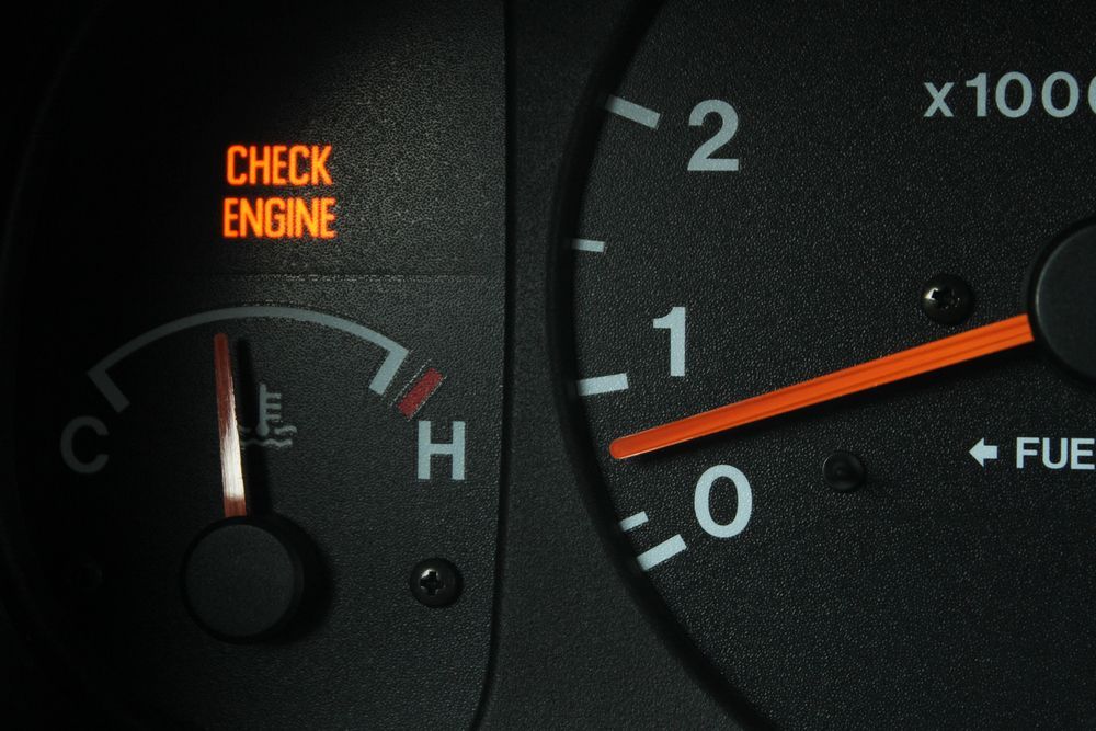 Car dashboard showing a lit check engine warning and gauges, with the temperature needle near hot.