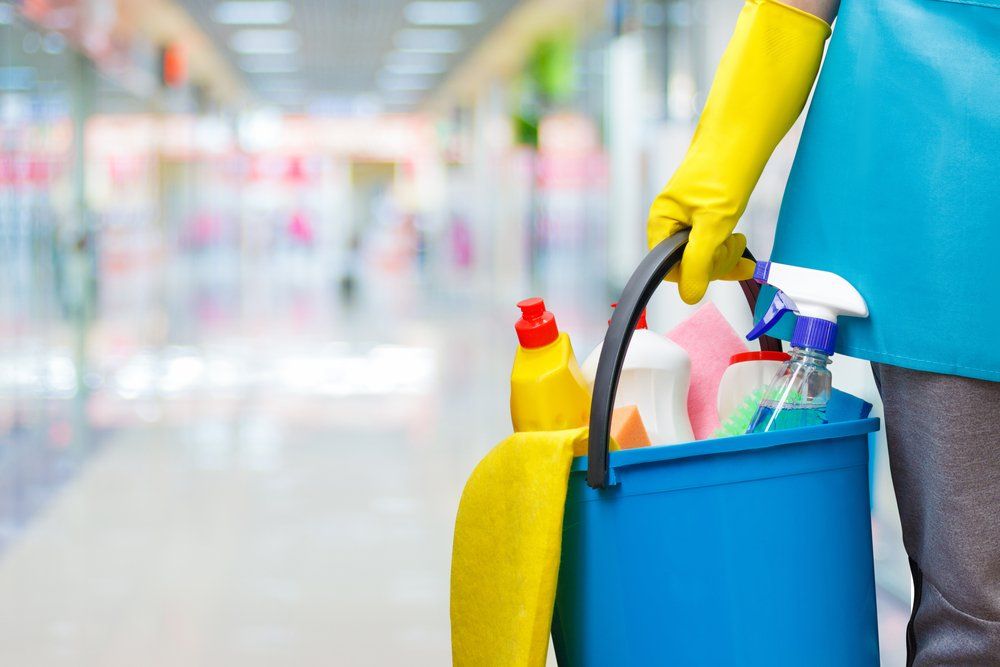 A woman in yellow gloves is holding a blue bucket filled with cleaning supplies.