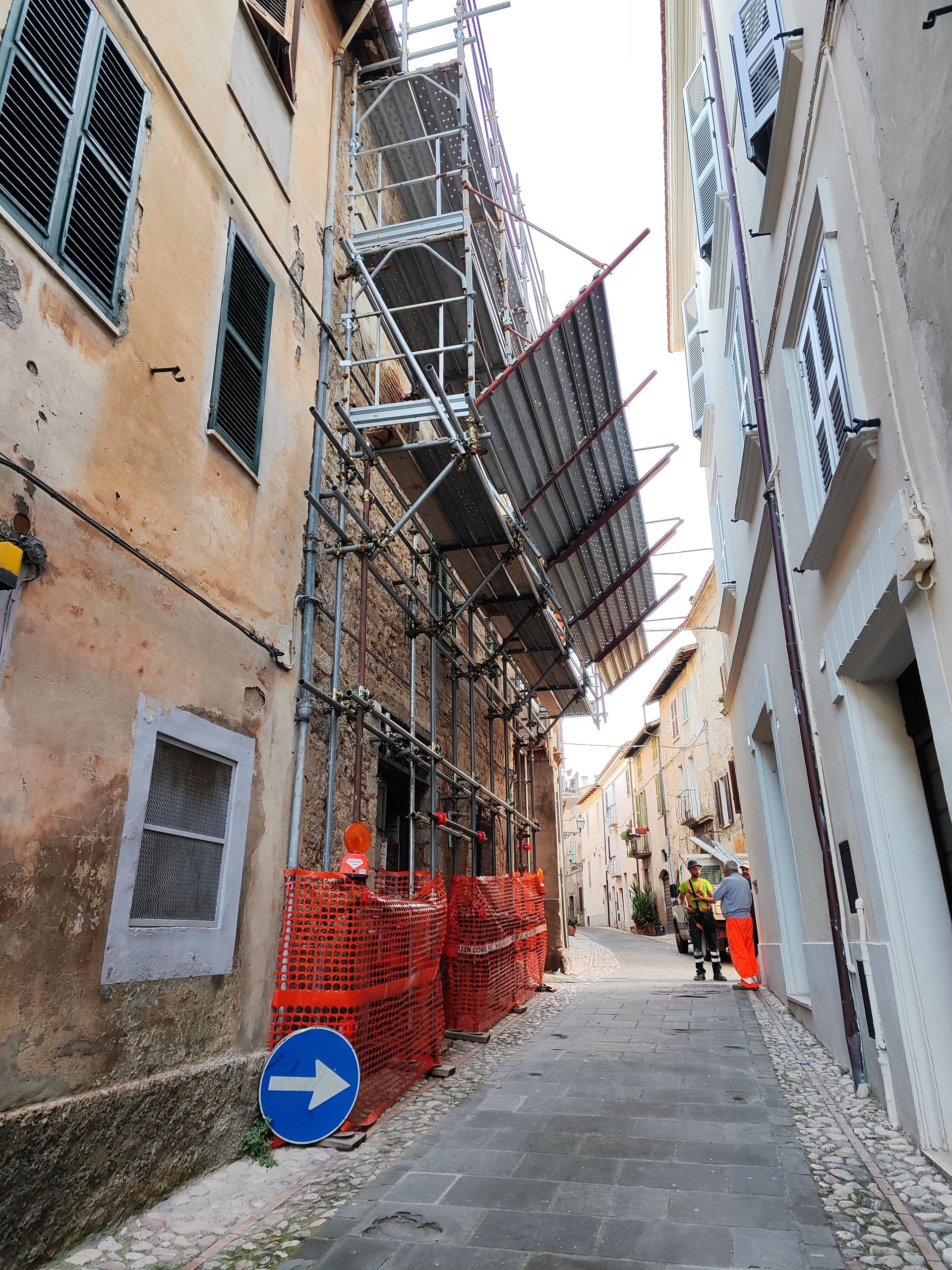 Strada stretta con impalcature sull'edificio a sinistra. Operai edili in lontananza.