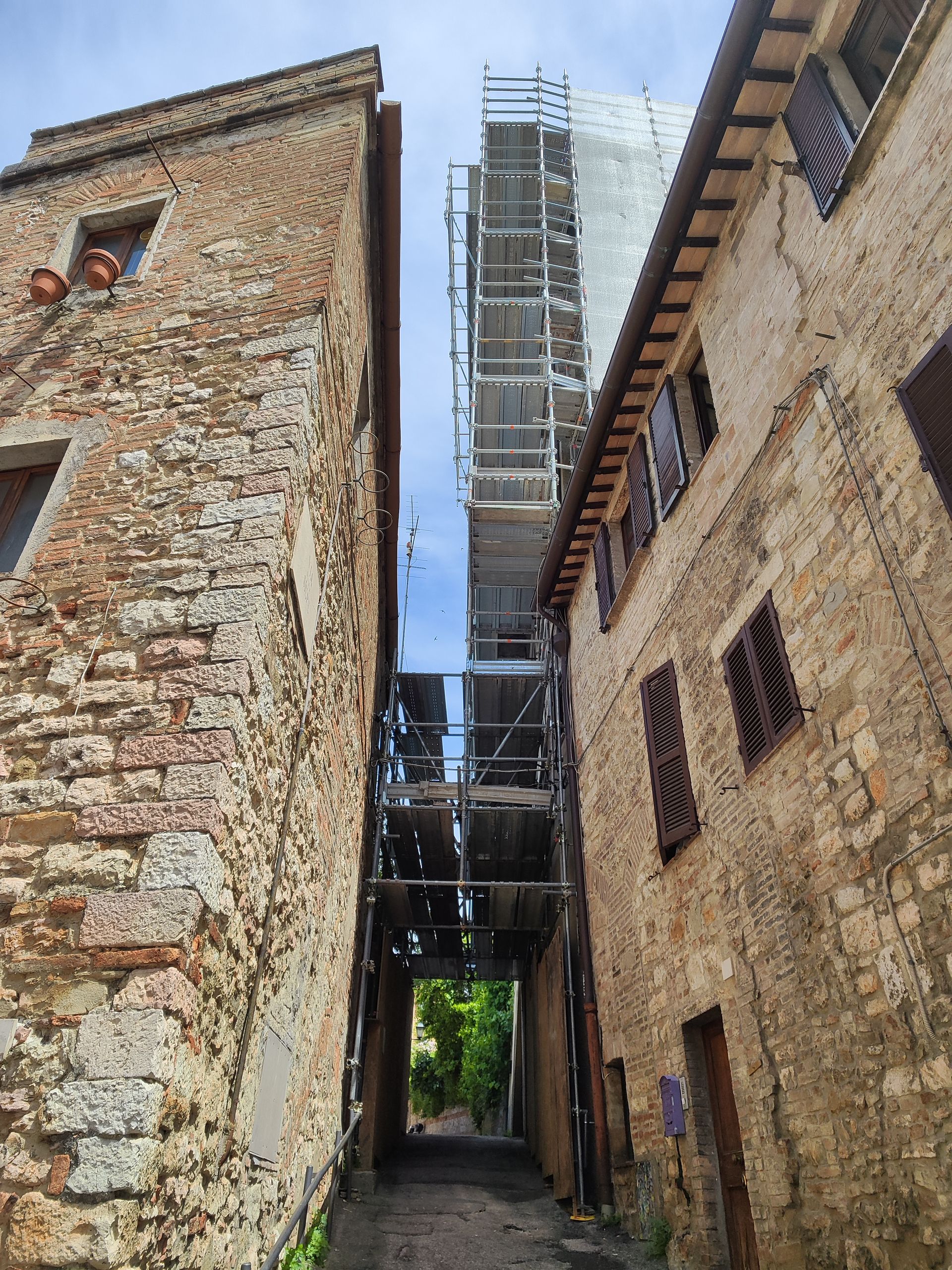 Stretto vicolo in pietra tra gli edifici, con impalcature che si protendono verso l'alto; in alto si vede il cielo azzurro.