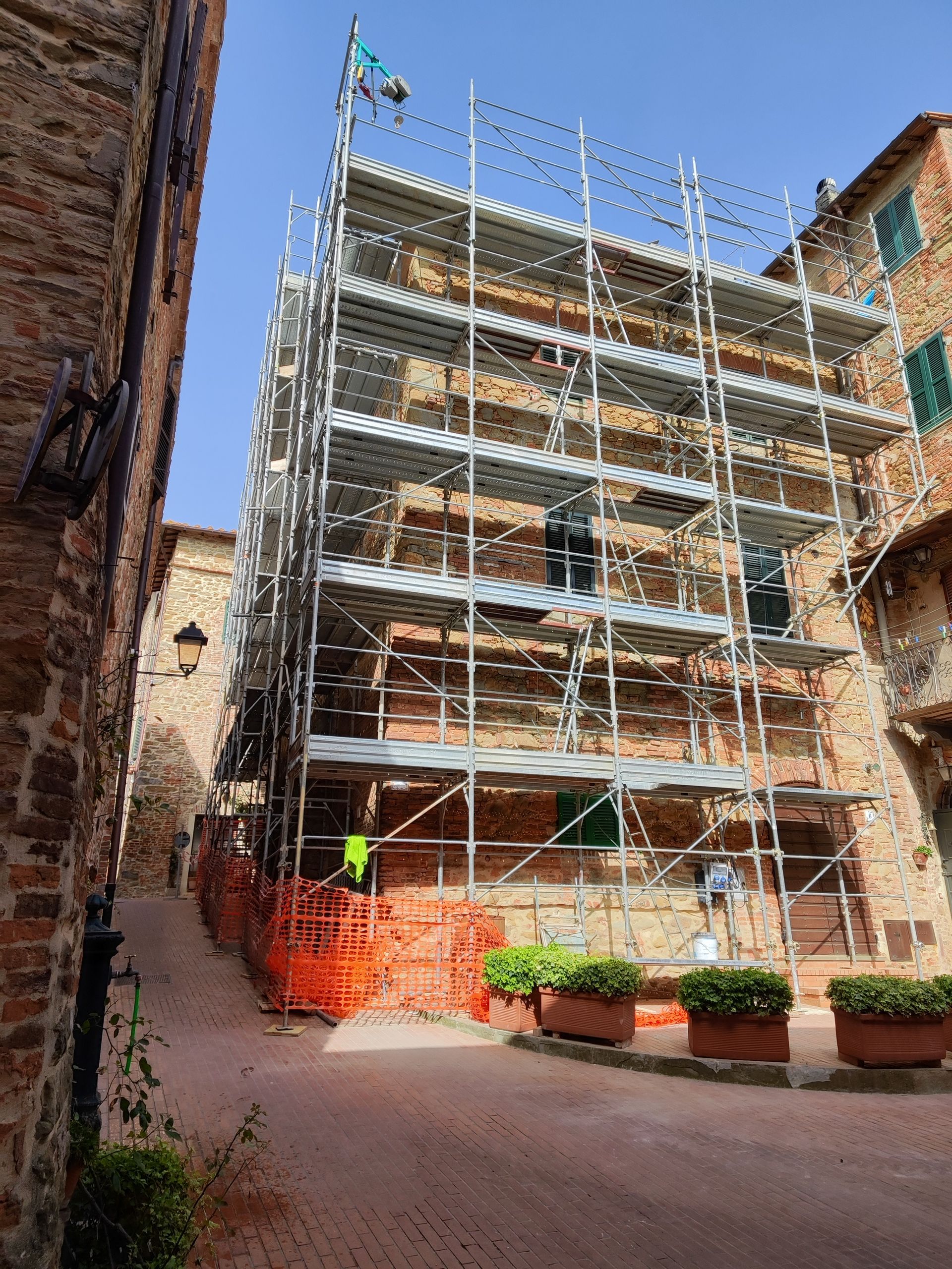 Un'impalcatura circonda un edificio in mattoni in una strada stretta. Sono visibili una recinzione di sicurezza arancione e piante in vaso.