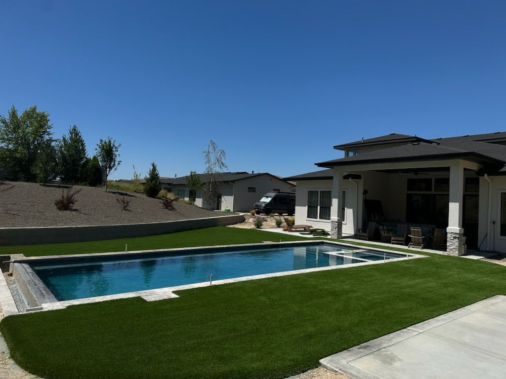 Backyard with rectangular pool, artificial turf, and a house with a covered patio under a bright blue sky.