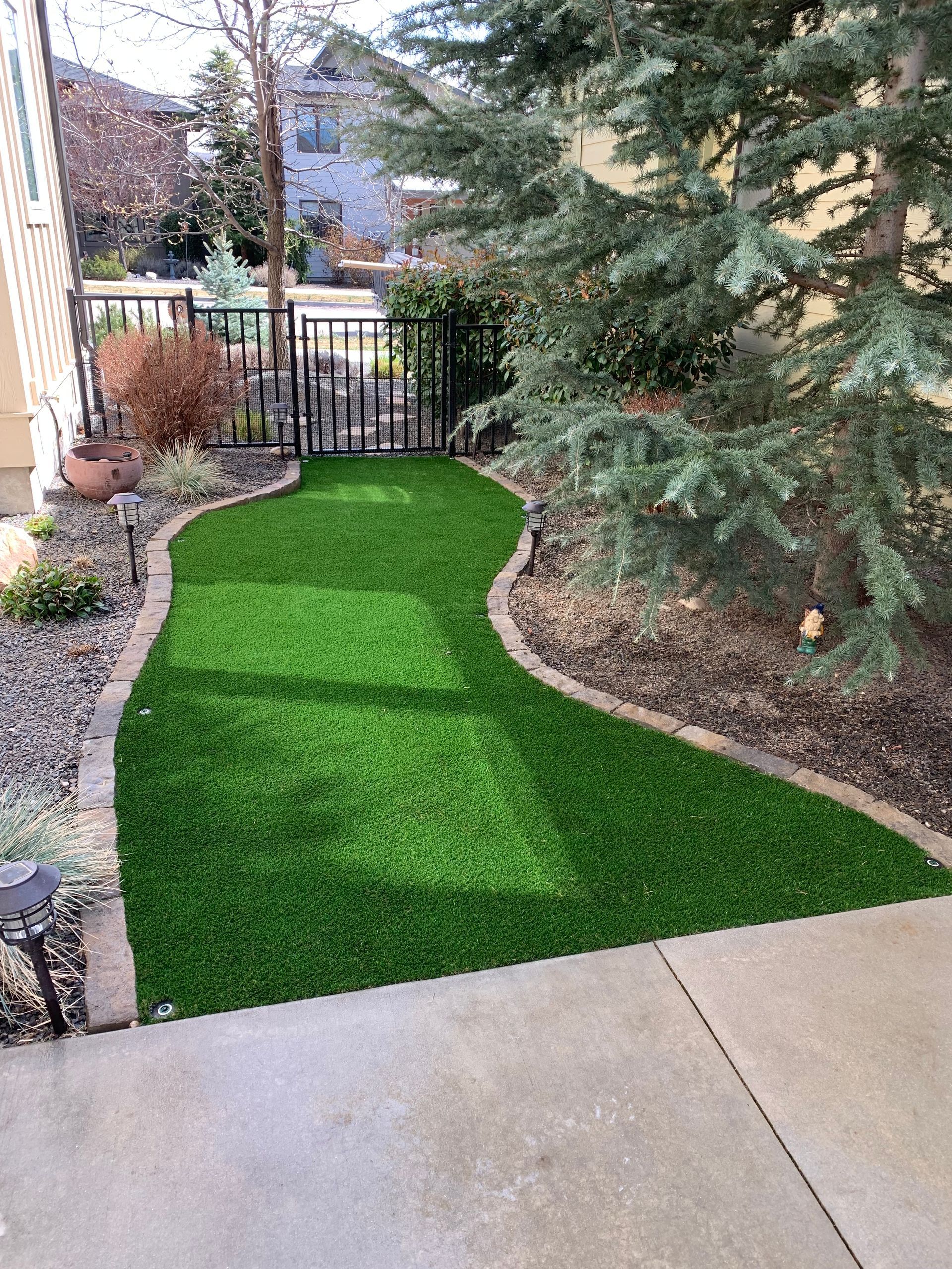 Small, artificial turf pathway with a curved border, next to a concrete patio and plants.