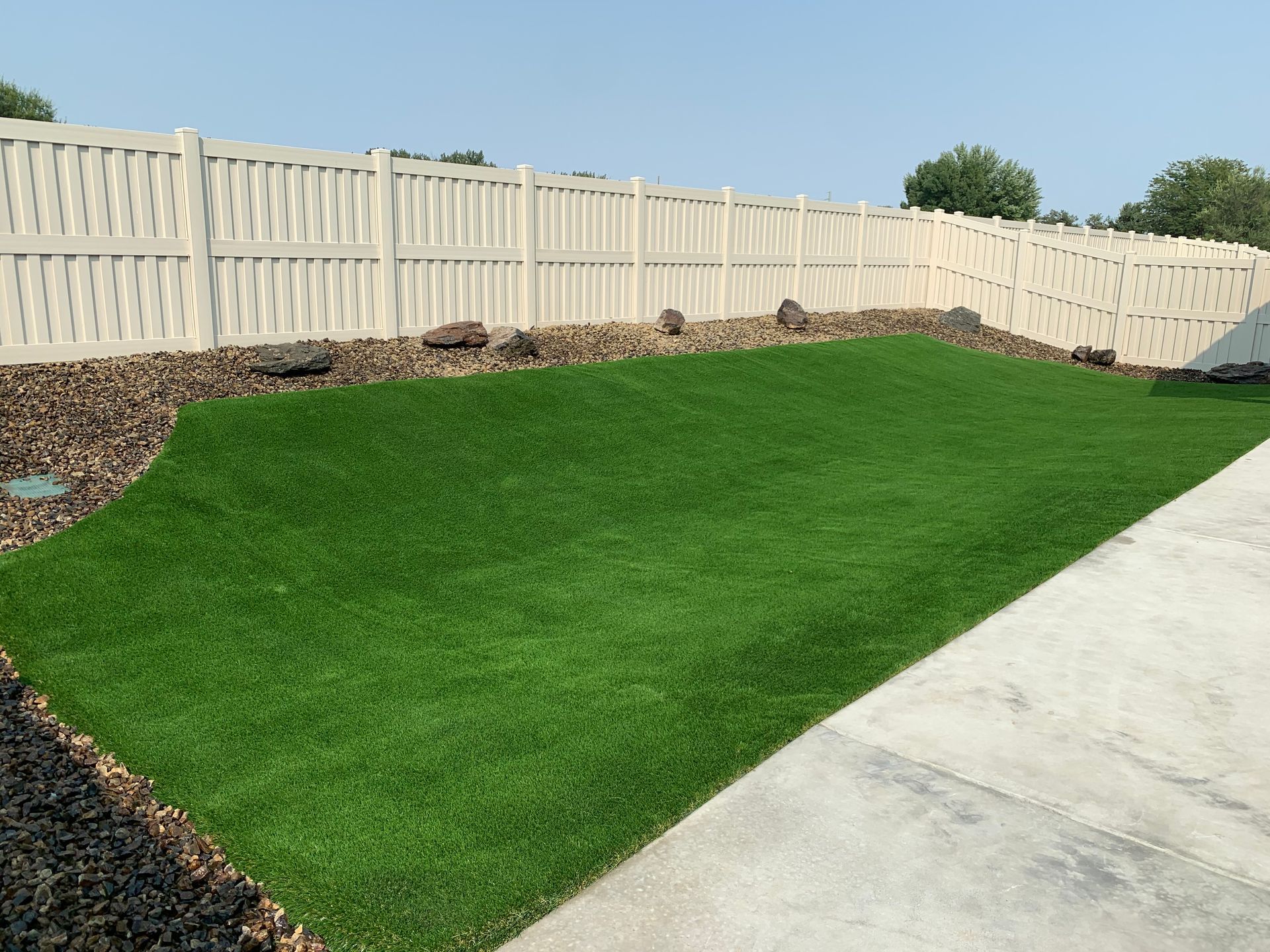 Green artificial turf yard bordered by dark rocks and a white fence.