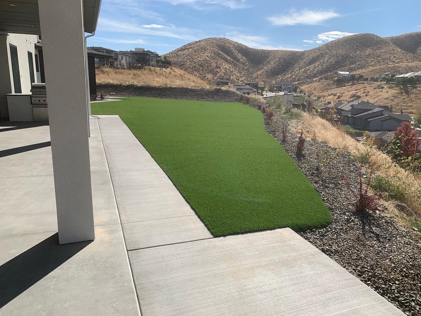 Backyard with artificial turf, concrete patio, and hillside view under a blue sky.