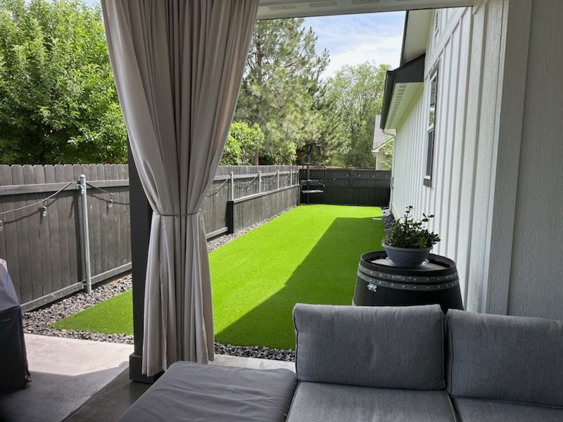 Outdoor patio with gray couch, curtains, artificial turf, and a wooden barrel with plants.