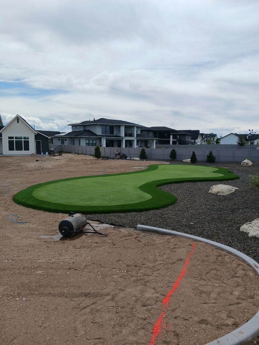 Green putting green with surrounding rock and dirt, near houses under cloudy sky.